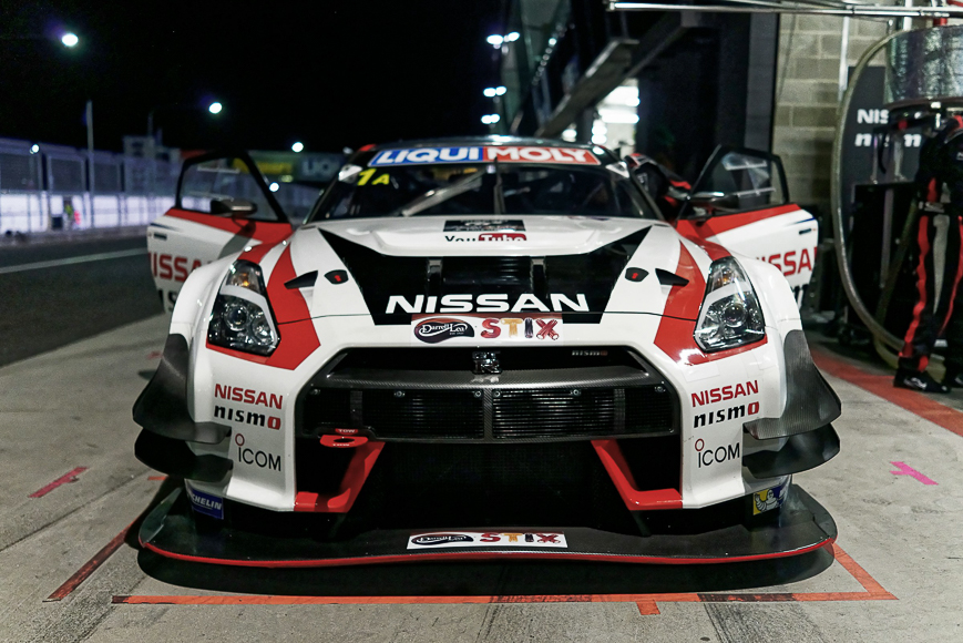 A Nissan GT-R NISMO race car with open doors is parked in a pit garage at night during a motorsport event.