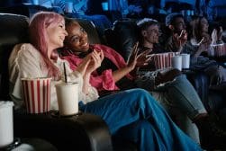 People sitting in a theater with relaxed seating, drinking and holding popcorn, clapping and enjoying the show.