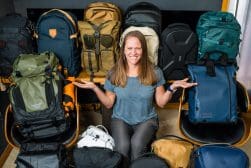 A person seated, smiling, surrounded by an array of different backpacks in various colors and styles.