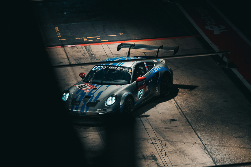A grey and blue race car with a large rear wing is parked in a pit lane under artificial lighting, viewed from above and partially obscured by shadow.