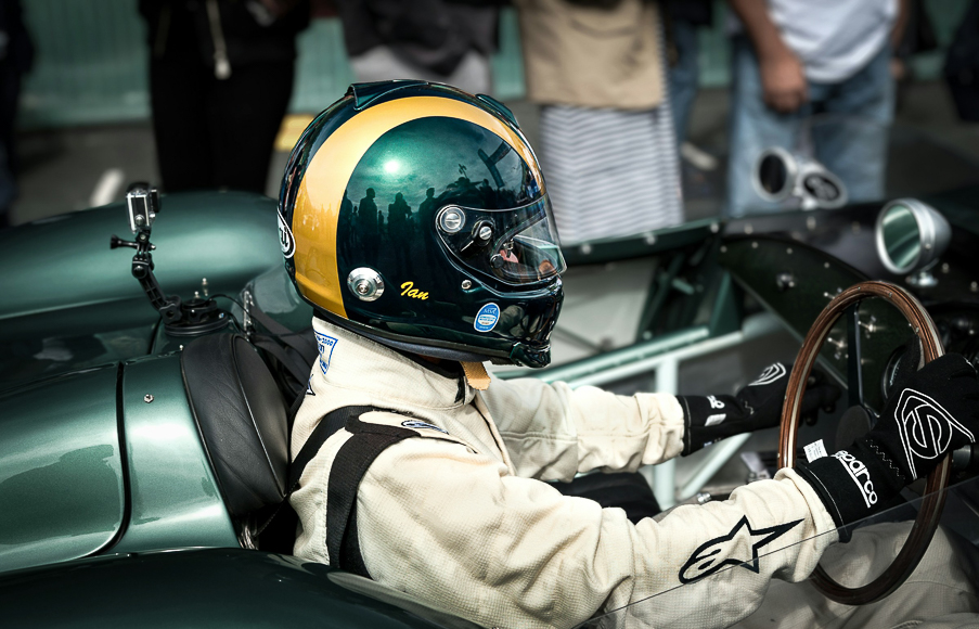 A driver in a cream racing suit and green helmet sits in a vintage green race car, holding the wooden steering wheel, with people standing in the background.