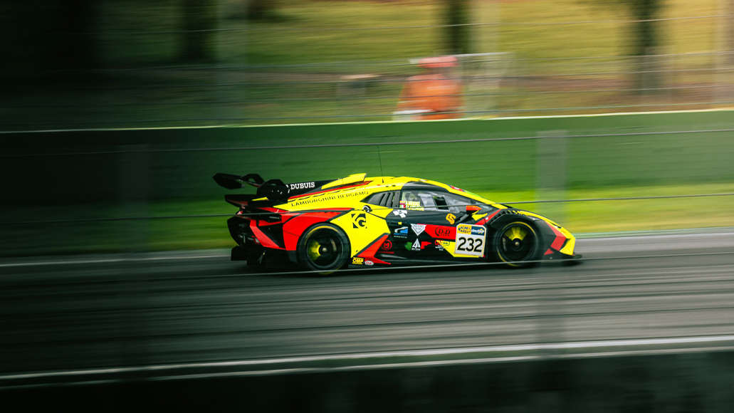 A yellow and black race car with number 232 speeds along a racetrack, with motion blur visible in the background and on the wheels.