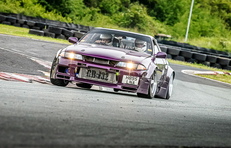 A purple sports car drifts around a track corner, with two helmeted drivers inside and tire barriers and greenery in the background.