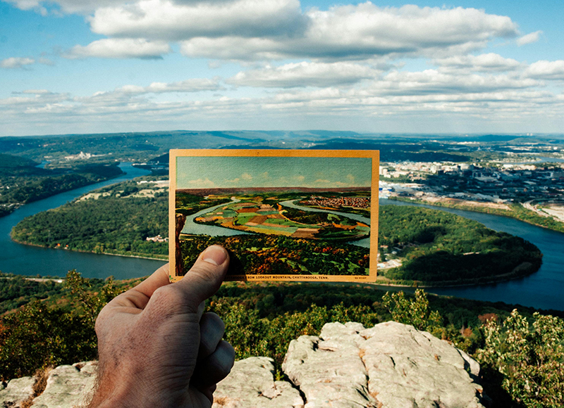 A hand holds a postcard showing a landscape that matches the real view behind it, overlooking a river, trees, and distant town under a cloudy sky.