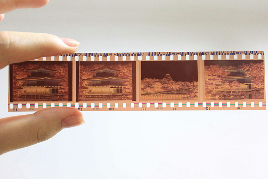 A hand holds a strip of photographic film negatives showing four images of a traditional building and landscape.