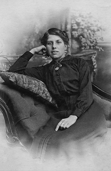 A woman in a dark dress sits on a cushioned chair, resting her head on her hand, with a neutral expression and a blurred background featuring flowers.