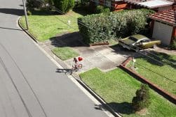 A person wearing a red hoodie rides a bicycle on a suburban street. A vintage car is parked in a driveway, and neatly trimmed lawns line the sidewalk.