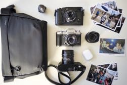 A camera bag, three cameras, two lenses, wireless earbuds, a lens cap, and several printed photos are arranged on a white surface.
