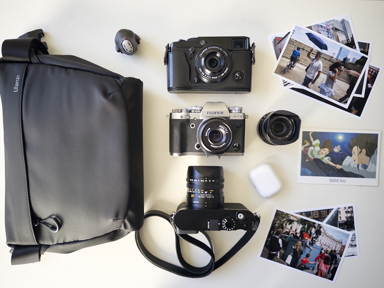 A camera bag, three cameras, two lenses, wireless earbuds, a lens cap, and several printed photos are arranged on a white surface.