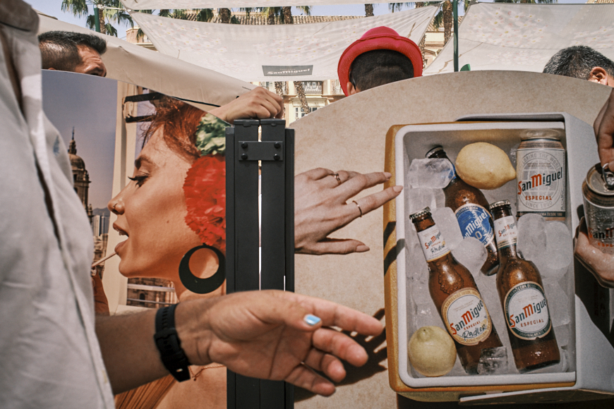 People stand behind a display showing a woman’s face and a cooler with San Miguel beers on ice. A hand with a cigarette is visible in the foreground.