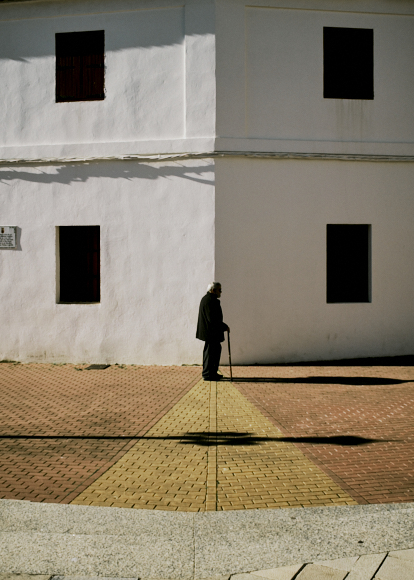 An elderly person with a cane stands on a patterned sidewalk at the corner of a white building with closed windows.