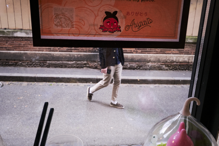A person walks on a street outside, partially obscured by a window sign with cartoon art and Japanese text. Dining items and utensils are visible inside by the window.