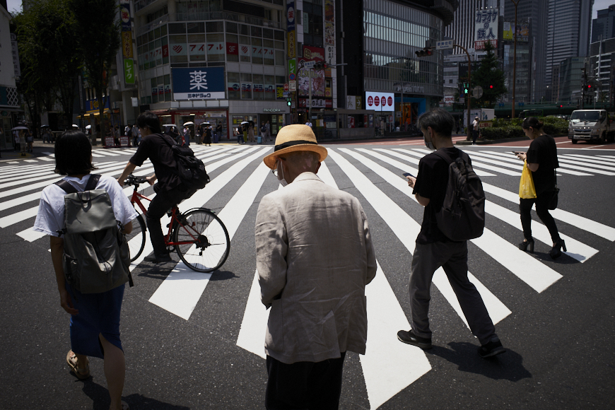 People walk and cycle across a large, striped crosswalk in a busy city area, with buildings and signage visible in the background.