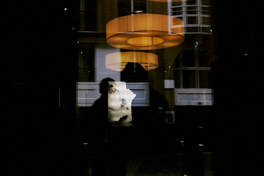 A person’s silhouette is reflected in a glass window, with round ceiling lights and a building’s facade visible in the background. A small poster is also reflected in the glass.