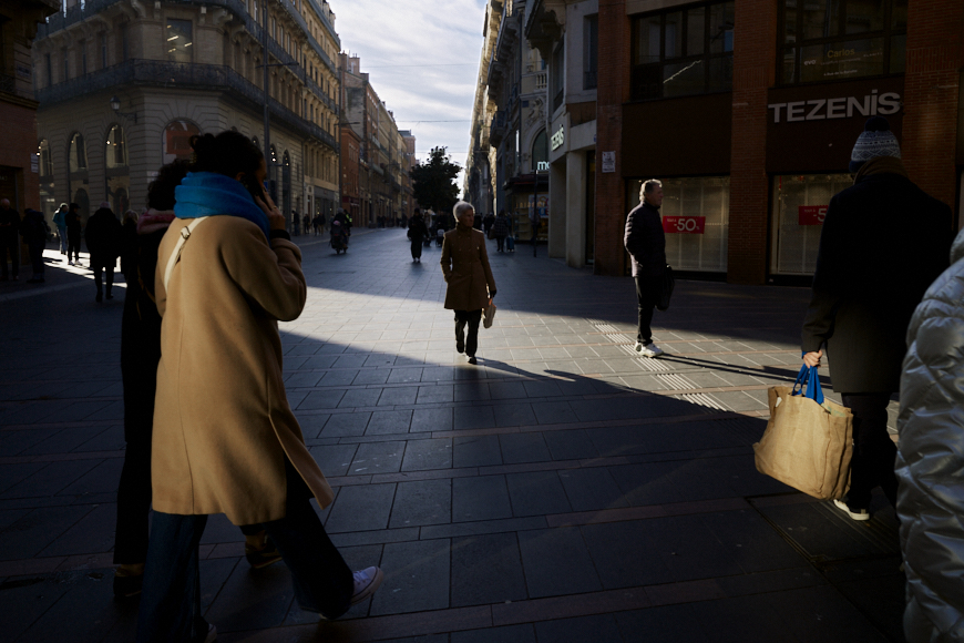 People walk along a sunlit city street, with some areas in shadow and others brightly illuminated. Several storefronts are visible in the background.
