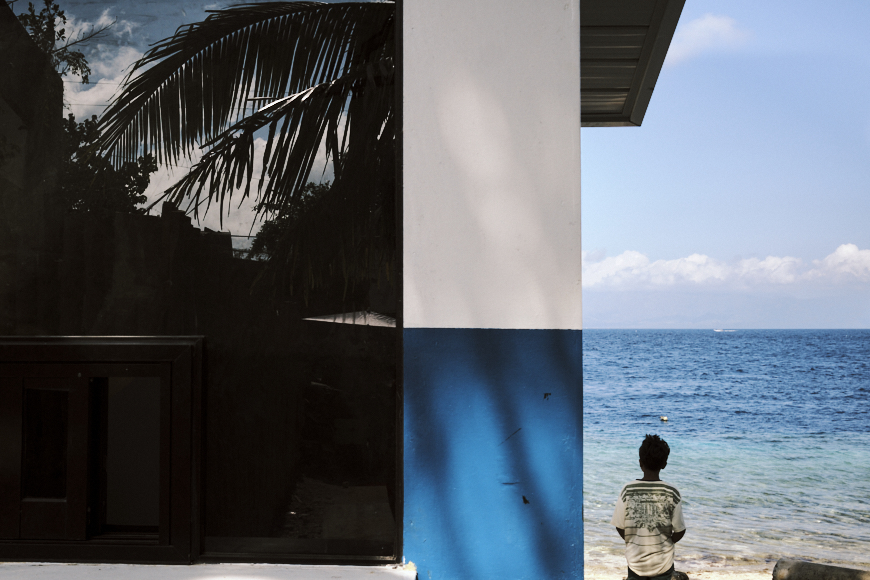 A person stands by the edge of a building with white and blue walls, looking out at the ocean; a palm tree is reflected in a nearby window.