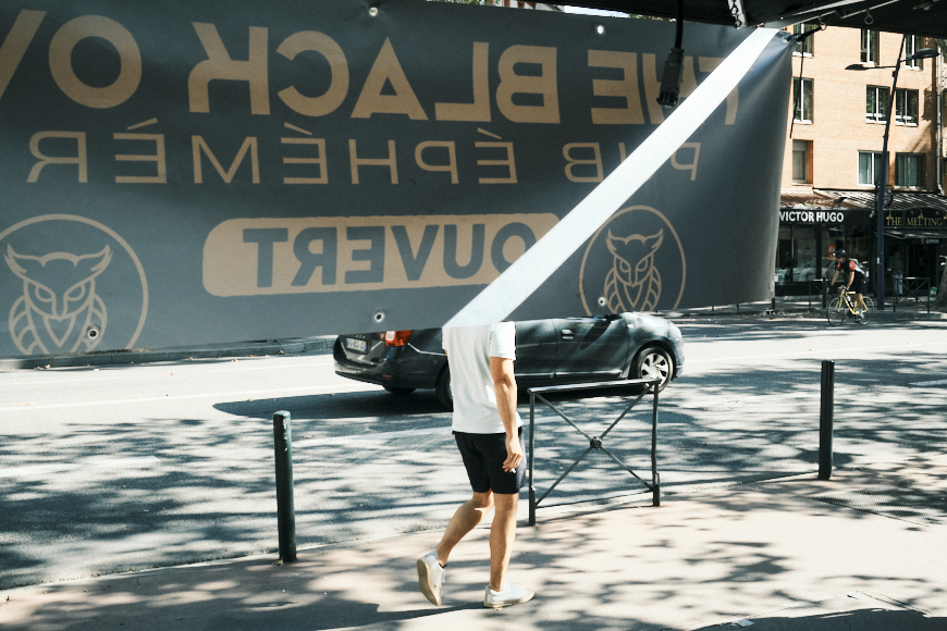 A person in shorts and a t-shirt walks behind a partially lowered banner on a sunny street, with cars and buildings visible in the background.
