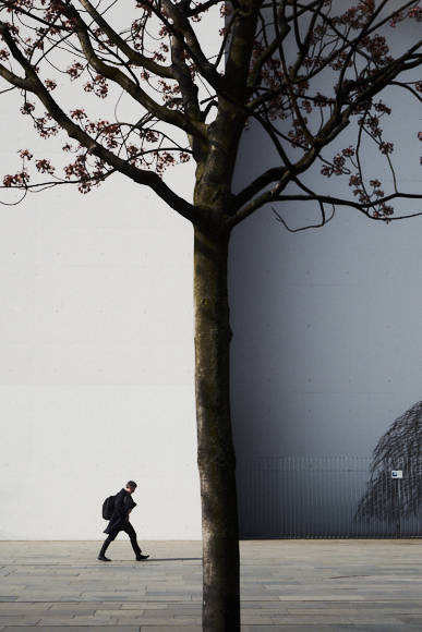 A person with a backpack walks on a paved sidewalk past a leafless tree in front of a large white wall.