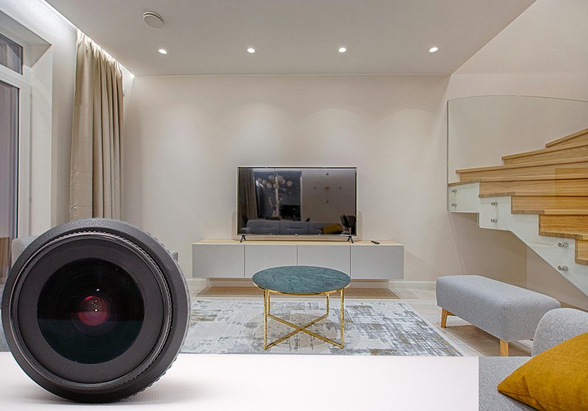 A modern living room with a large flat-screen TV on a white entertainment unit, a round coffee table with a green top, and a staircase with a wooden finish. A camera lens is in the foreground. A modern living room with a large flat-screen TV on a white entertainment unit, a round coffee table with a green top, and a staircase with a wooden finish. A camera lens is in the foreground.