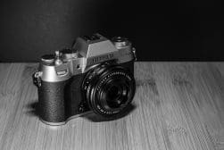 Black and white image of a Fujifilm X-T3 camera on a wooden surface, featuring a Fujinon aspherical lens.