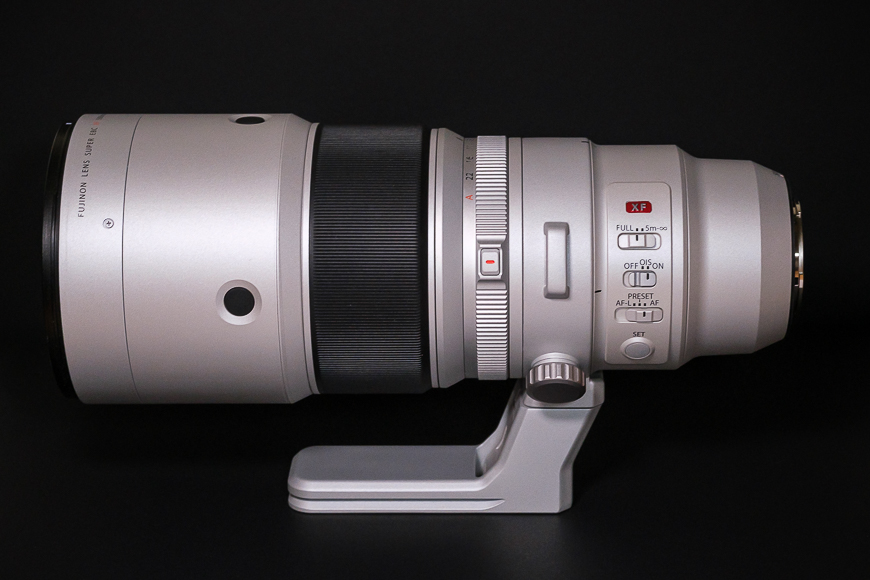A large silver camera lens with a black focus ring and various control switches is mounted on a tripod foot against a dark background.