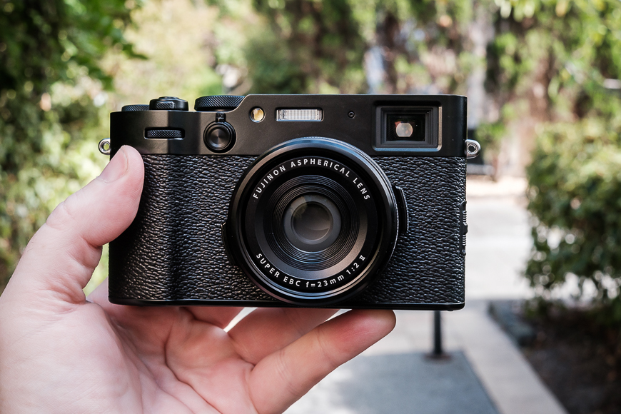 Hand holding a black rangefinder-style camera with a prime lens outdoors.