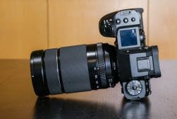 A DSLR camera with a zoom lens attached, resting on its side on a dark surface, against a wooden background.