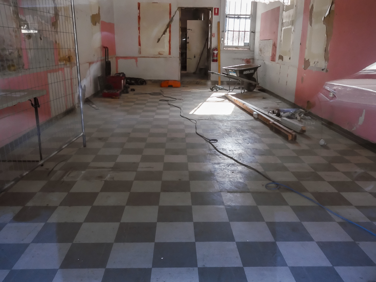 A room with checkered flooring, peeling paint, construction tools, and materials scattered around, suggesting ongoing renovation work.