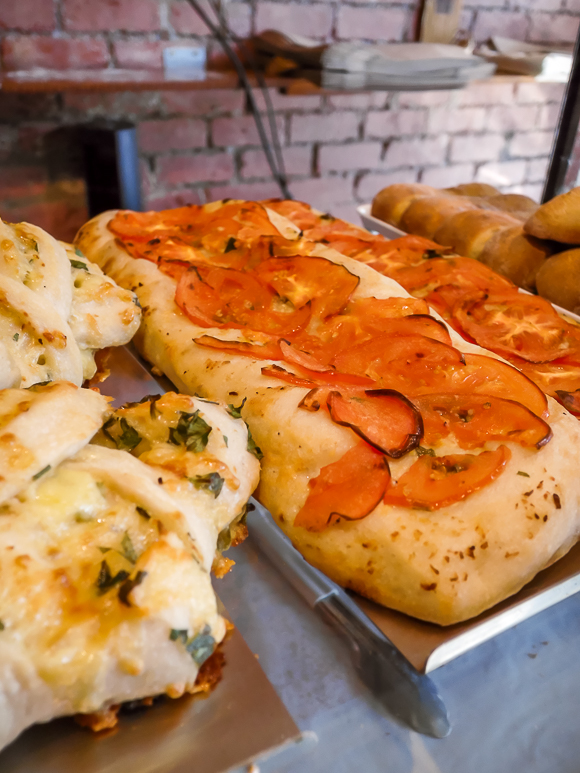 Close-up of two types of rectangular focaccia bread, one topped with sliced tomatoes and the other with cheese and herbs, displayed on metal trays in a bakery.