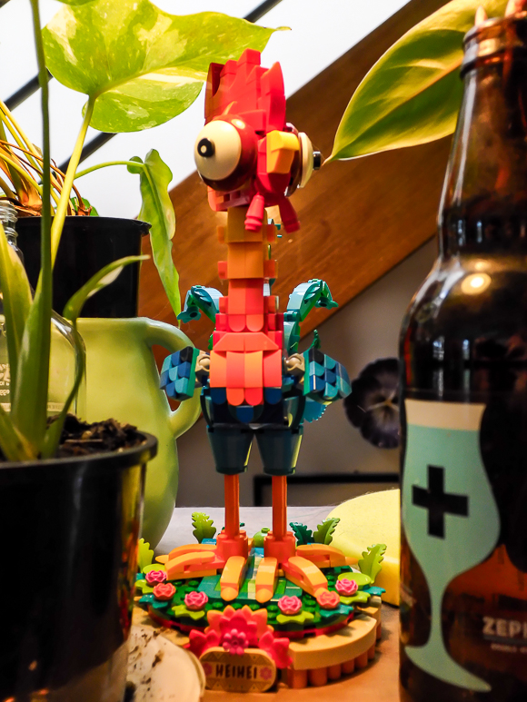 A colorful, block-style toy bird figure stands on a decorated base surrounded by potted plants and a bottle on a table.