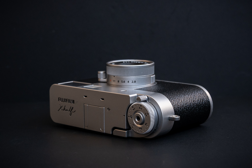 A silver and black Fujifilm X100V camera is displayed against a black background, shown from an angled top view.
