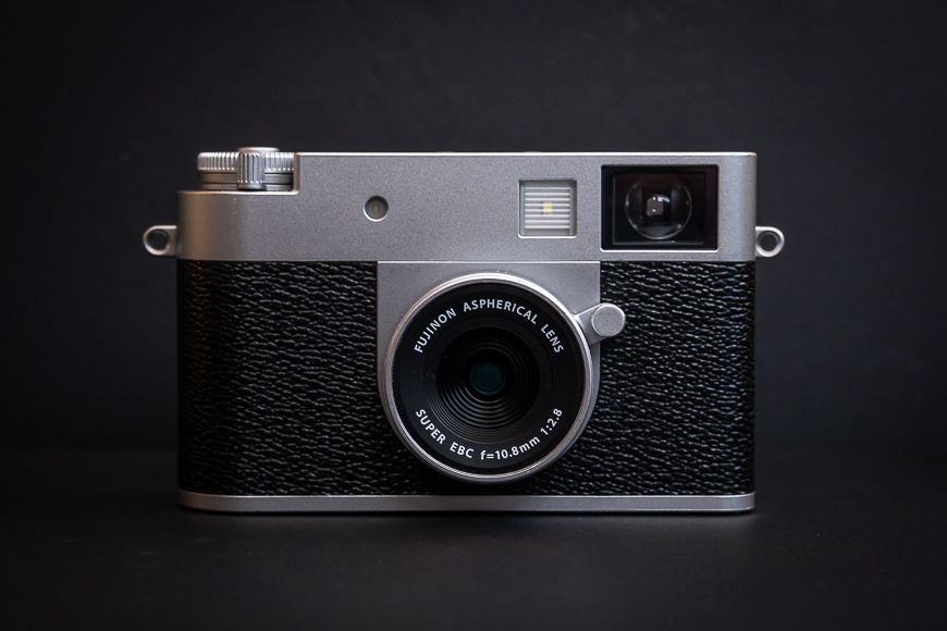 A silver and black Fujifilm camera with a Fujinon aspherical lens is placed against a plain black background.