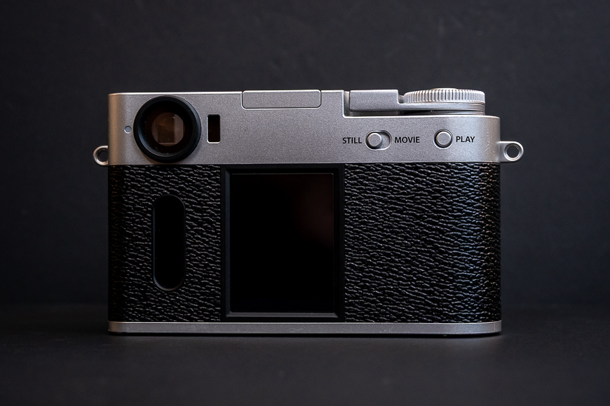 A digital camera with a black and silver body is shown from the back, featuring a blank screen, control buttons labeled "Still," "Movie," and "Play," and a textured grip.
