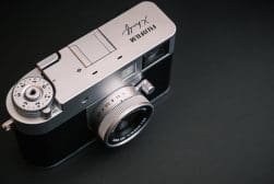 A silver Fujifilm X-series digital camera is placed on a dark surface, showing the top dials and lens.