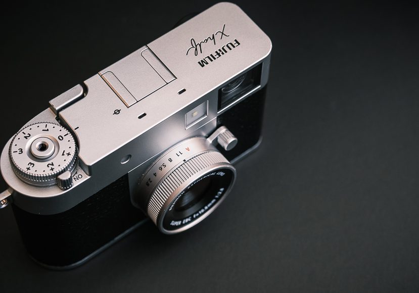 A silver Fujifilm X-series digital camera is placed on a dark surface, showing the top dials and lens. A silver Fujifilm X-series digital camera is placed on a dark surface, showing the top dials and lens.