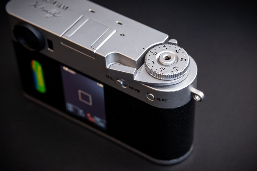 A close-up of the top rear section of a silver digital camera, showing its control dial, display screen, and textured grip, set against a dark background.