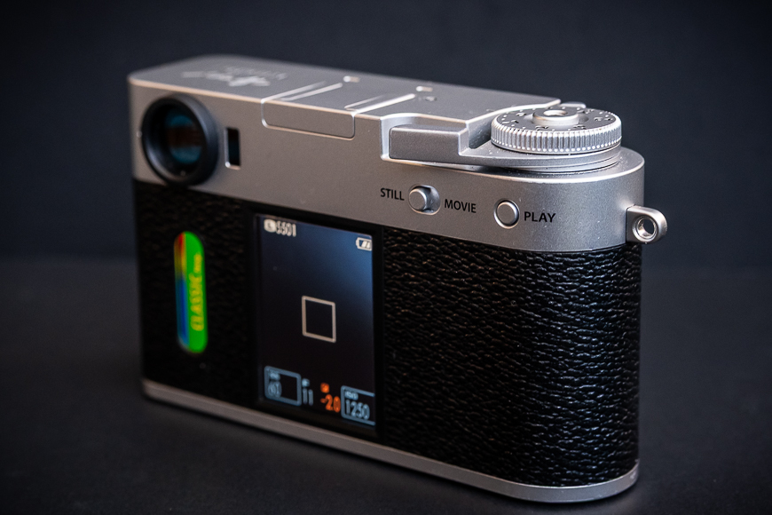 A digital camera with a textured black body, silver top, mode dial, and a display screen on the back, shown against a dark background.