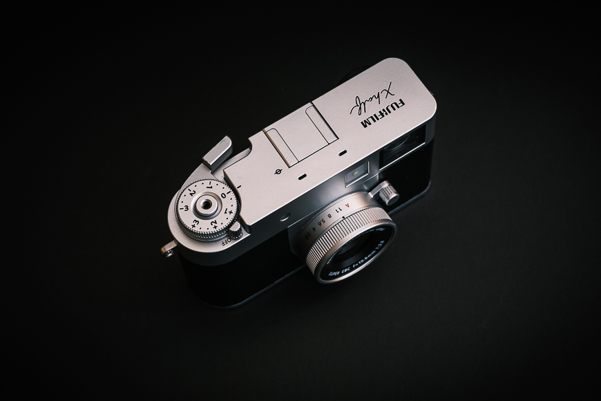 A silver and black Fujifilm X-series camera is shown from above against a dark background, highlighting the dials and lens.