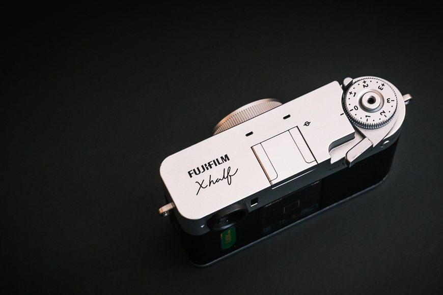 Top view of a Fujifilm X half camera showing the shutter speed dial, shutter button, and engraved branding on a dark background.