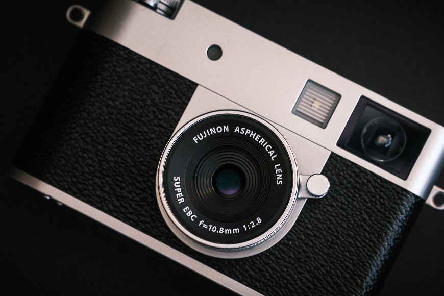 Close-up of a Fujifilm camera with an aspherical lens, showing lens details and textured body against a dark background.