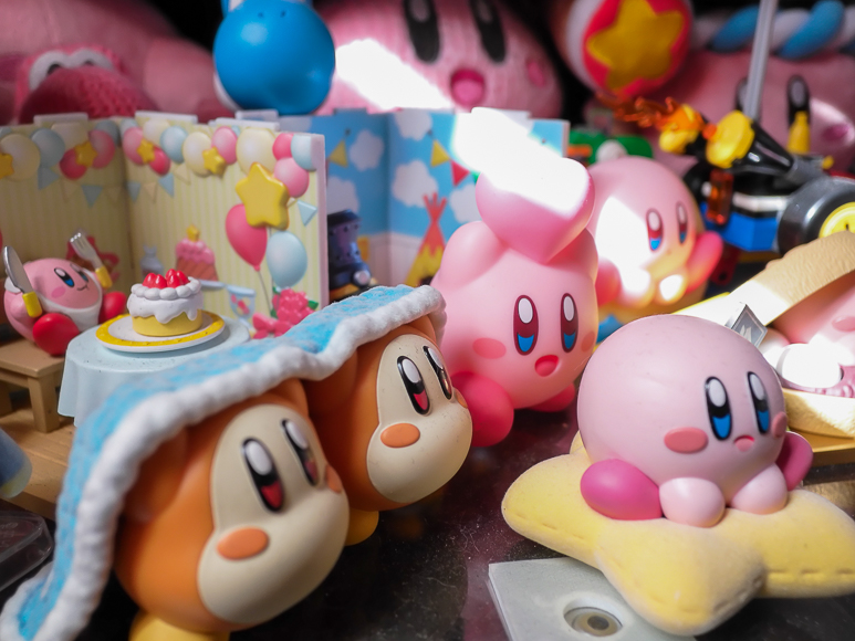 Assorted Kirby and Waddle Dee figurines are arranged on a surface, with a birthday-themed miniature scene in the background.