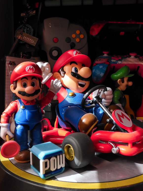Two Mario figurines, one standing and one driving a go-kart, are displayed next to a Luigi figure and a POW block toy with Nintendo controllers in the background.