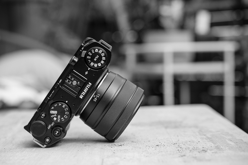 Black and white image of a Fujifilm camera with a lens, positioned on a wooden surface.
