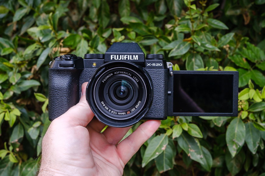 A person holding a fujifilm x-s20 camera with the screen flipped out against a backdrop of green leaves.