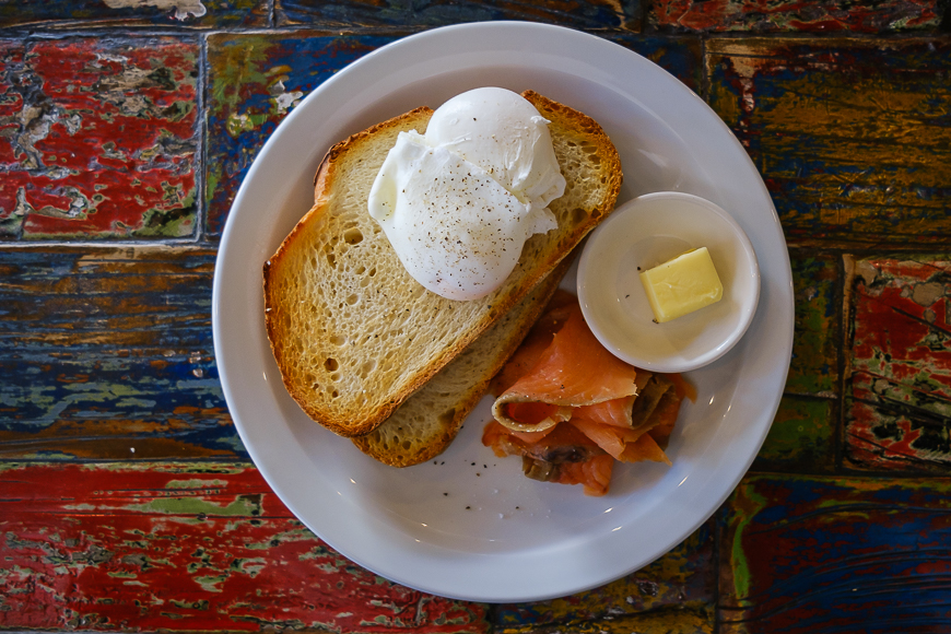 Poached eggs on toast with smoked salmon, accompanied by a pat of butter, served on a white plate over a colorful table.