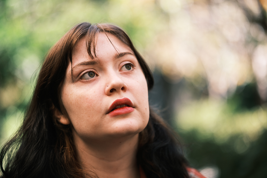 Woman looking upwards thoughtfully in a park, with soft, blurry green foliage in the background.