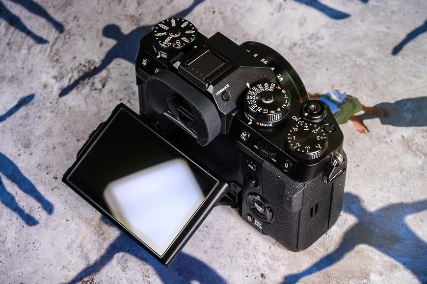 A digital camera with a tilting lcd screen and multiple control dials, resting on a textured surface.