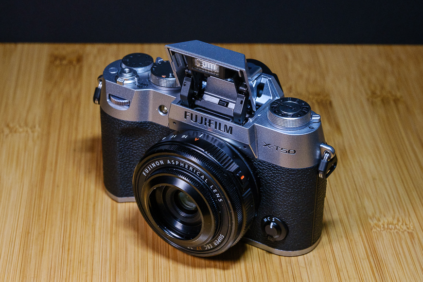 A Fujifilm X-T50 camera with a Fujinon Aspherical lens attached, set on a wooden surface, with the viewfinder partially opened.
