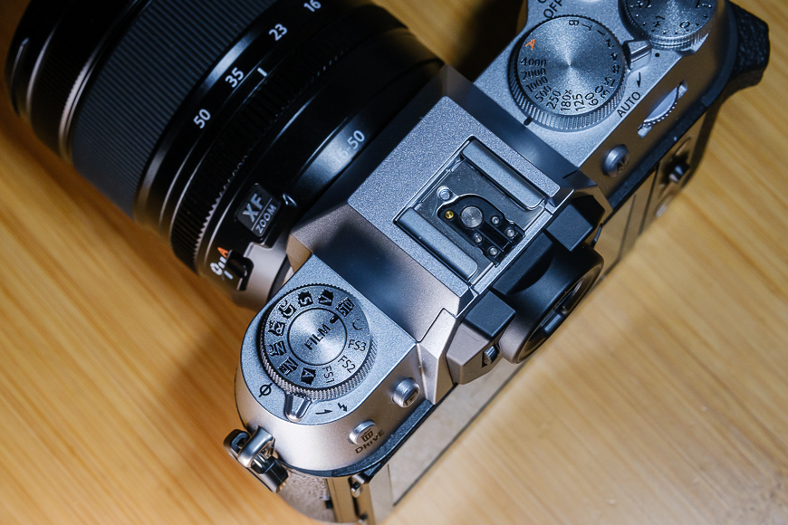 A top view of a digital camera with its various dials, buttons, and a lens attached, placed on a light wooden surface.
