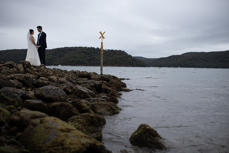 A bride and groom stand together on a rocky shore by a calm lake, with forested hills in the background under a cloudy sky.